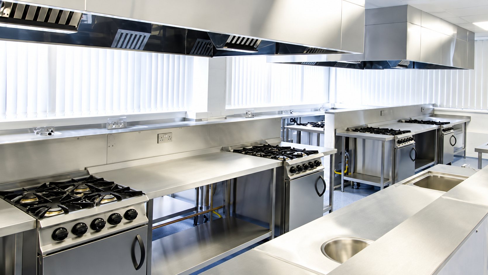 Intérieur d’une cuisine professionnelle moderne équipée de cuisinières à gaz, de plans de travail en inox et de hottes aspirantes.