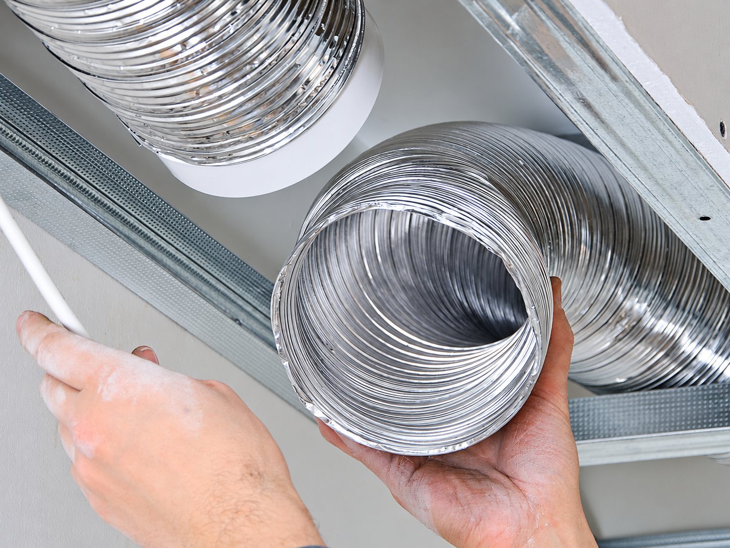 Deux mains d’ouvrier tiennent une gaine de ventilation flexible en aluminium installée entre les rails d’un faux plafond.